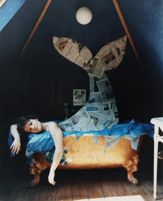 An unusual photograph depicts a person reclining in a bathtub beneath a whale tail sculpture made of newspaper inside a dark room.