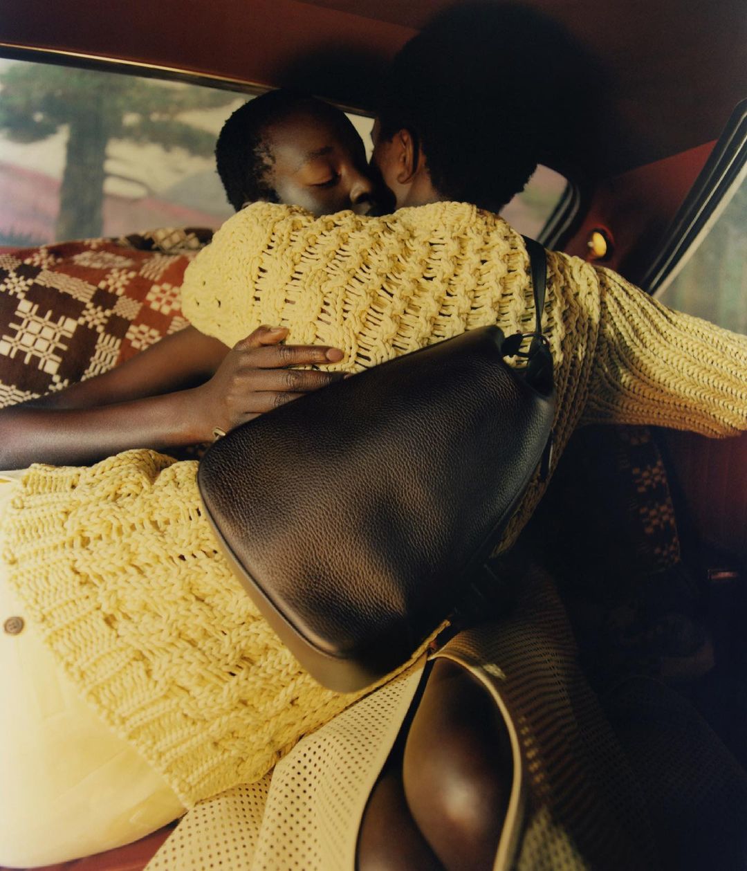 Two women embrace in car with a black handbag and light-yellow dress visible. Interior features patterned blanket and brown car interior.
