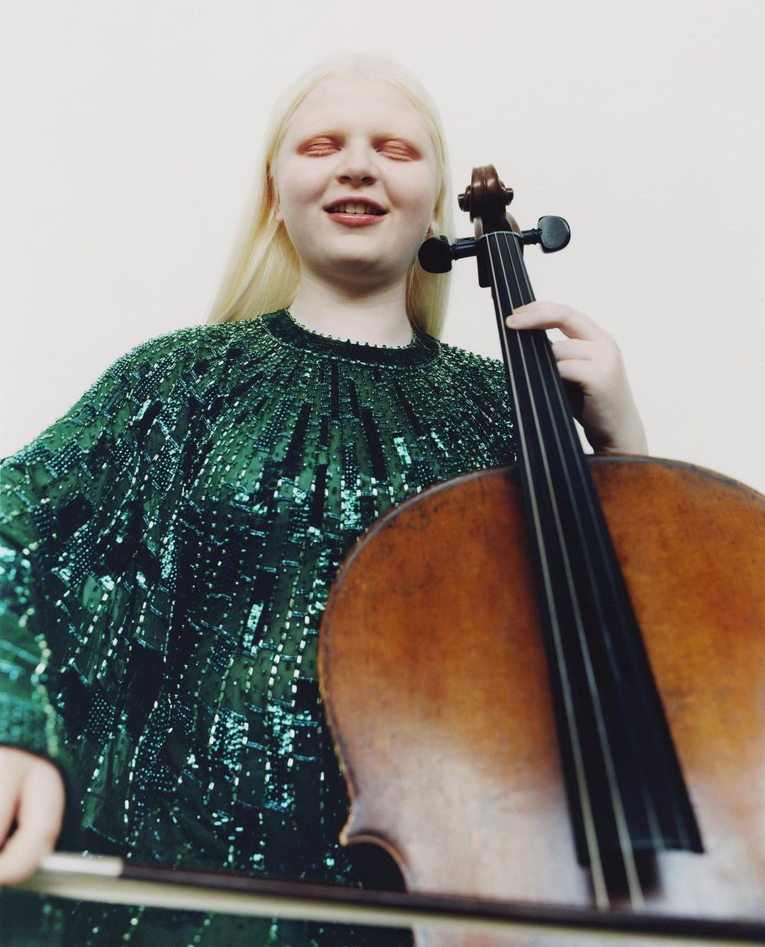 Albino woman playing cello in photo studio. She is wearing an emerald green beaded top with long blonde hair in a medium shot.