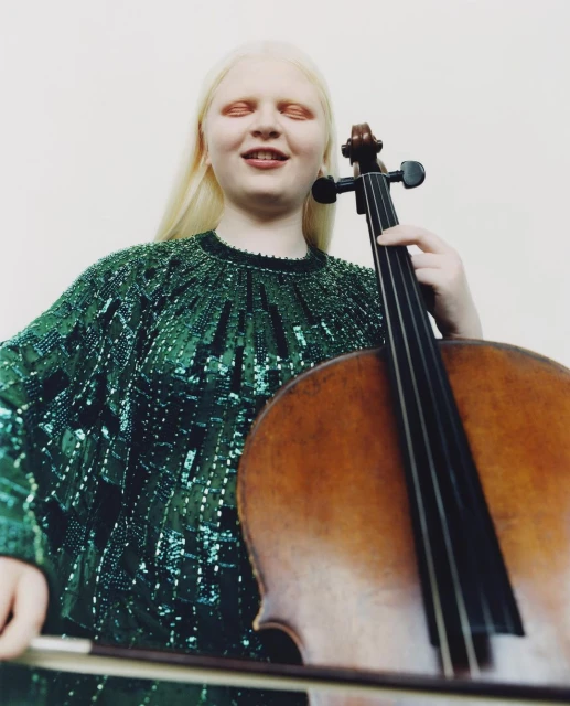 Albino woman playing cello in photo studio. She is wearing an emerald green beaded top with long blonde hair in a medium shot.