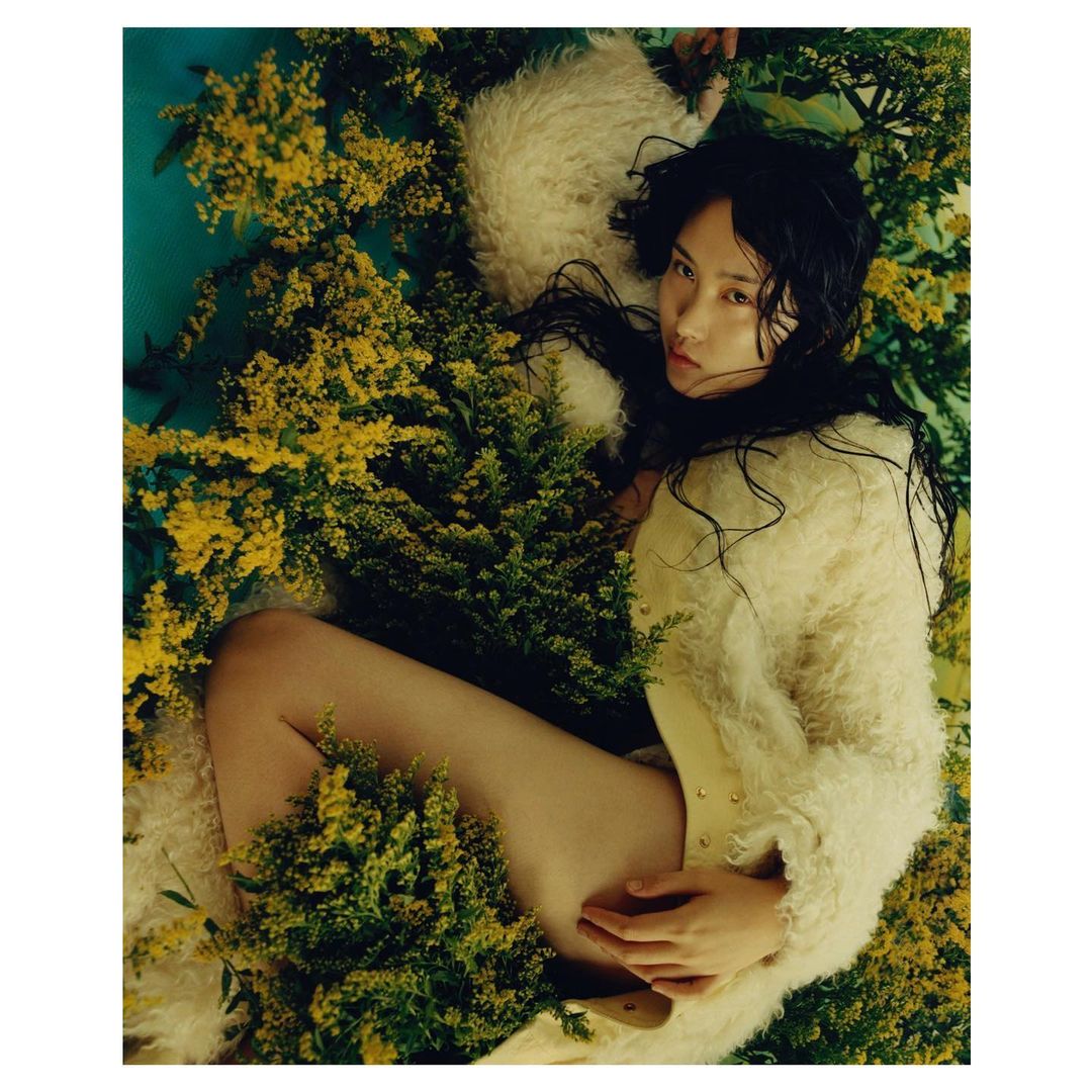 Woman in shaggy coat lying amongst goldenrod flowers; fashion editorial with nature elements, neutral tones and soft light.