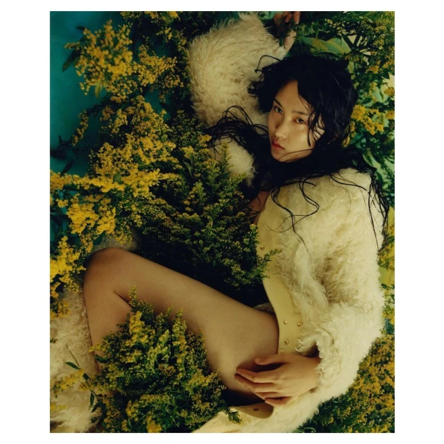 Woman in shaggy coat lying amongst goldenrod flowers; fashion editorial with nature elements, neutral tones and soft light.