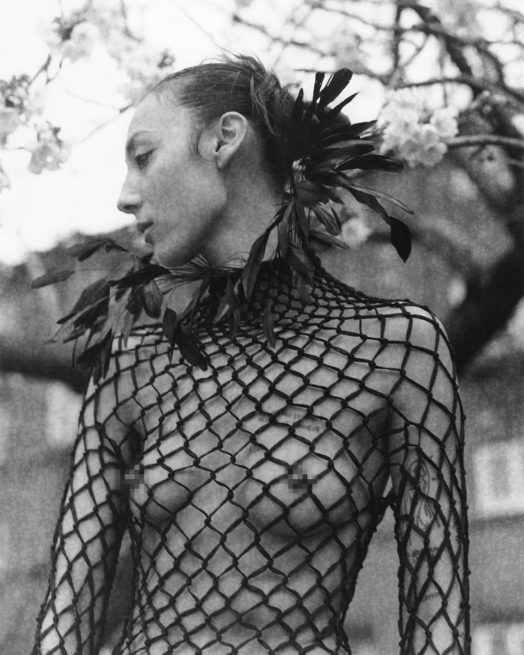 Monochrome portrait of a slender adult woman in a netted top with feathered details, complemented by cherry blossoms in the background.