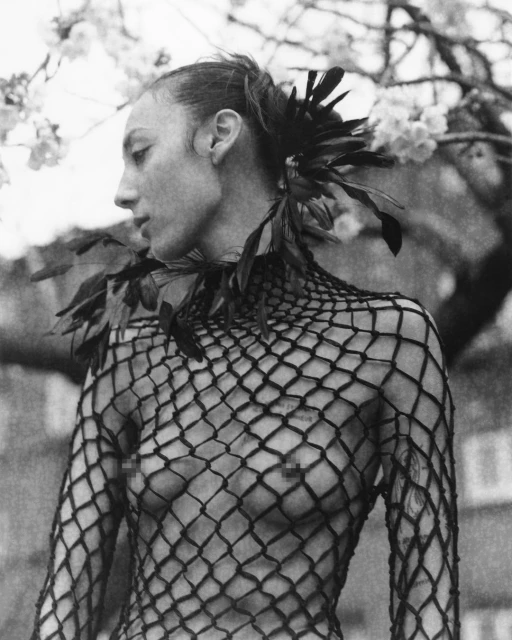 Monochrome portrait of a slender adult woman in a netted top with feathered details, complemented by cherry blossoms in the background.