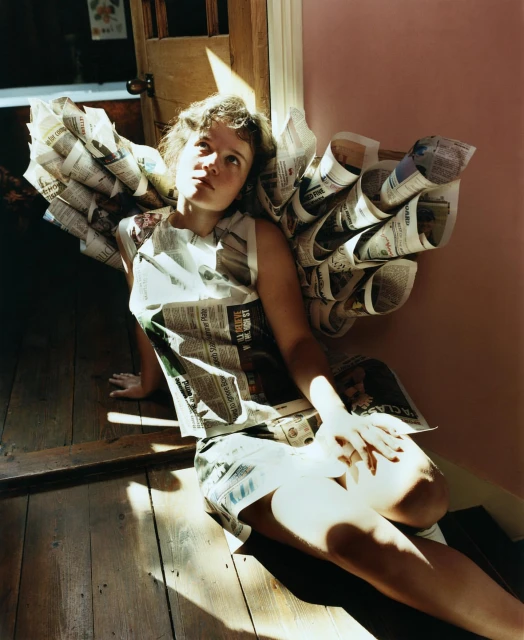 Young woman seated indoors, wearing a newspaper dress and newspaper wings in bright sunlight with a thoughtful expression.