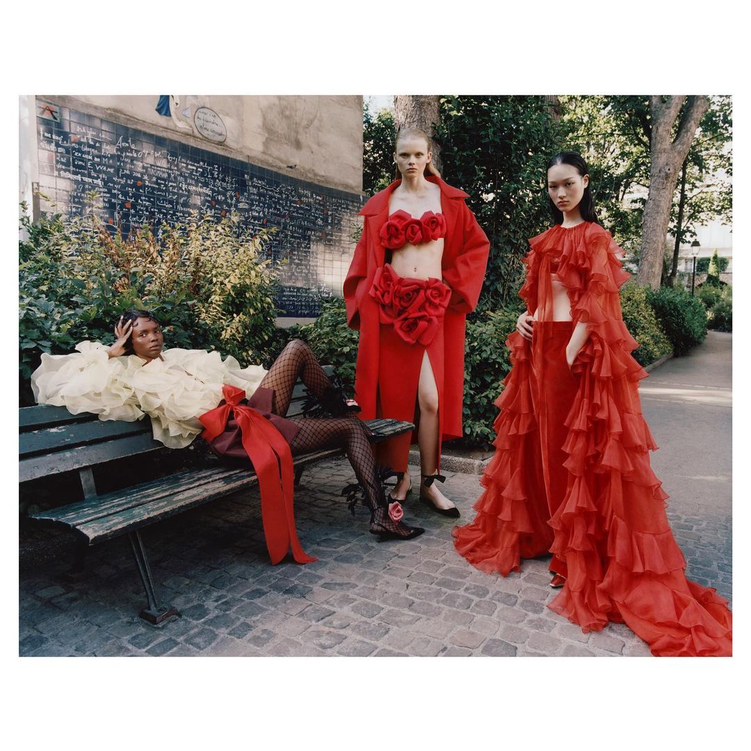 Three female fashion models pose in red and yellow couture outfits on a park bench in an outdoor setting with trees and a wall.