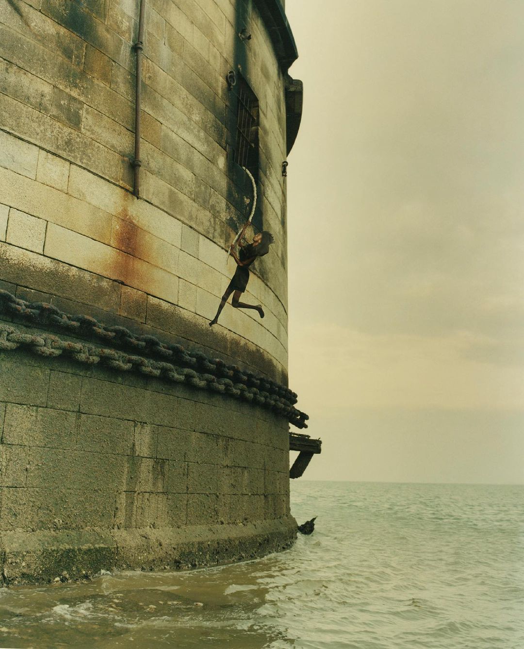 Figure rappelling down the exterior stone wall of a coastal fortress above choppy water