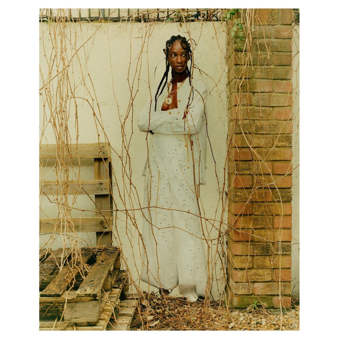 A Black woman in a white outfit stands with arms crossed against a cream wall, framed by vines and a brick column in an outdoor setting.