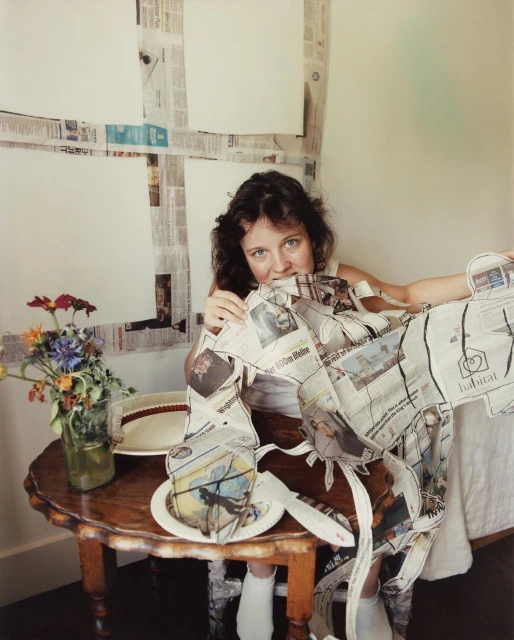 Woman in paper-covered outfit sits at table with flowers, plates, and utensils. Background is a wall decorated with newspaper.