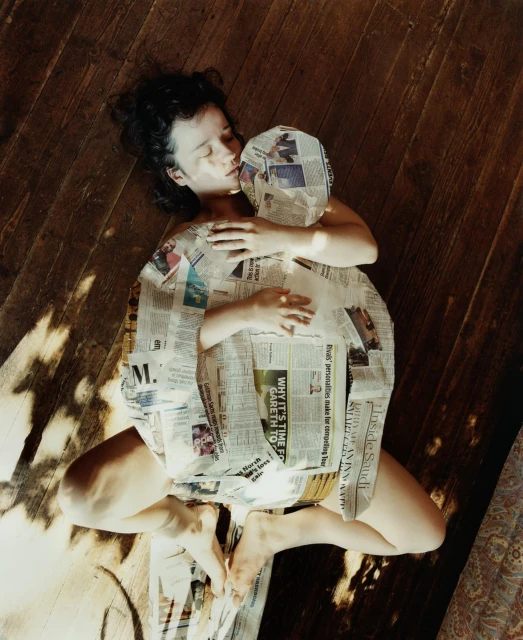 Woman lies on wooden floor embracing a newspaper-covered form in sunlit room, creating a surreal, intimate, and artistic composition.
