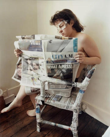 Young woman reading newspaper while sitting on a chair covered in newspaper in an indoor setting with wooden floor.