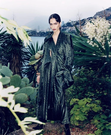 Model in dark green patterned coat standing outdoors amidst lush tropical foliage and ocean backdrop
