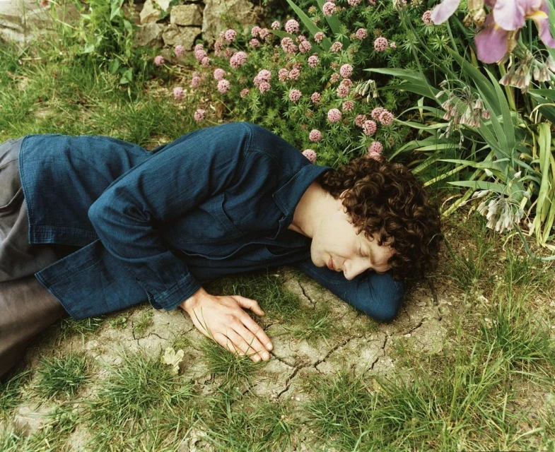 Adult male with curly hair rests in a grassy outdoor environment with pink flowers, wearing a dark-blue button-down shirt.