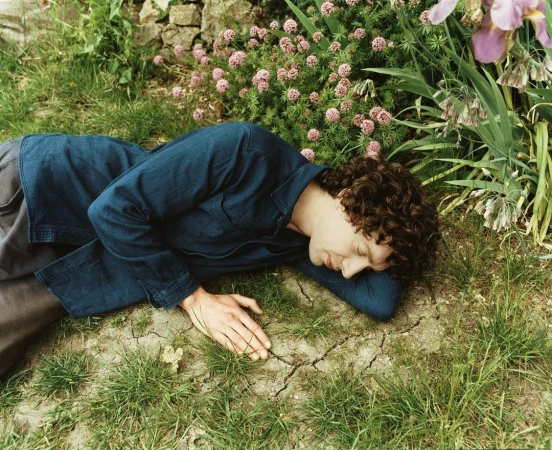 Adult male with curly hair rests in a grassy outdoor environment with pink flowers, wearing a dark-blue button-down shirt.