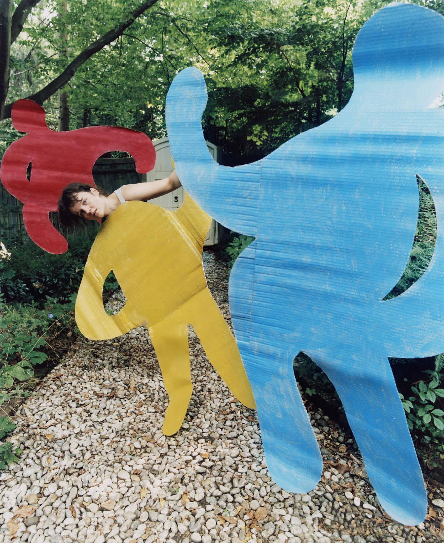 Outdoor shot of a woman behind blue, yellow, and red man-shaped, wooden art pieces, with a gravel path and lush green trees.