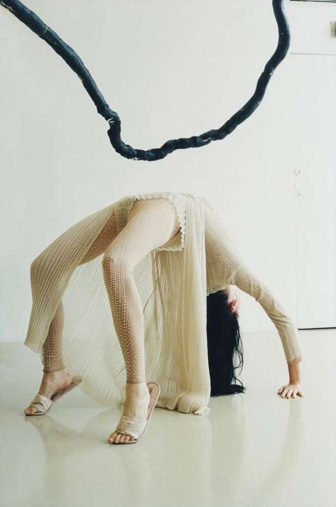 Woman in sheer dress does backbend under dark hanging sculpture in studio. Cream colors and minimalist, airy studio lighting.