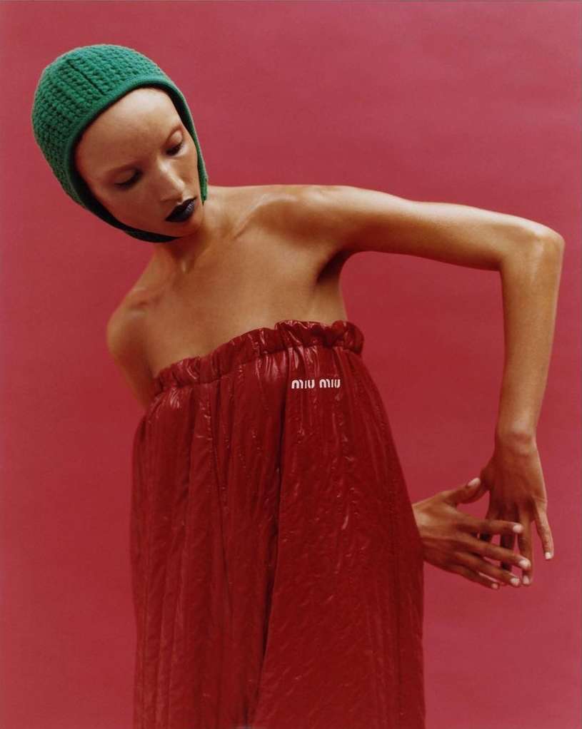 Fashion shot of a woman in a red dress and green knit hat against a pink backdrop. Dark lipstick and shiny skin add to the aesthetic.