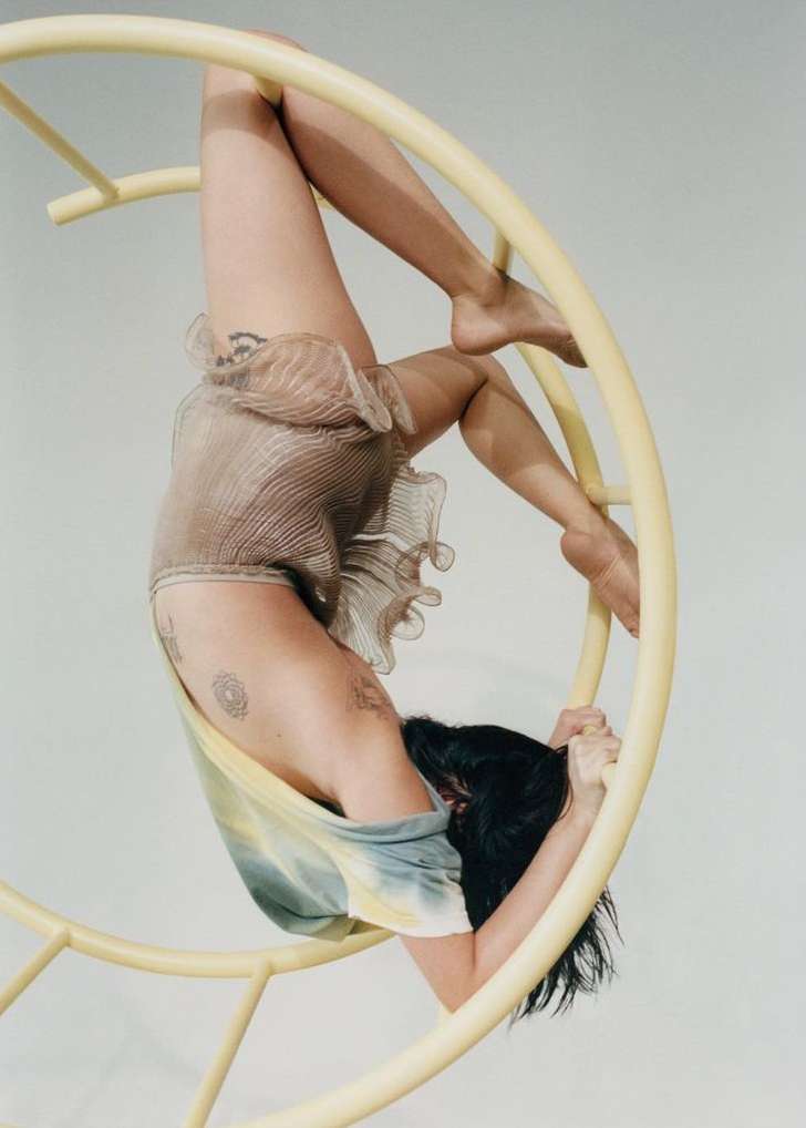 A barefoot woman is hanging upside down in a yellow circular apparatus in a studio, wearing a ruffled beige bodysuit and a tie-dye shirt.