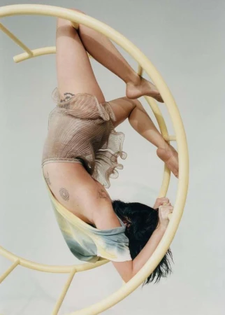 A barefoot woman is hanging upside down in a yellow circular apparatus in a studio, wearing a ruffled beige bodysuit and a tie-dye shirt.