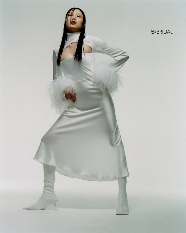Asian woman poses in a white high-neck dress with feathered sleeves and matching boots against a pale background in a fashion shot.