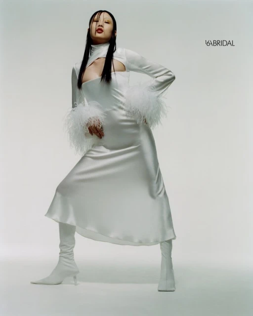 Asian woman poses in a white high-neck dress with feathered sleeves and matching boots against a pale background in a fashion shot.
