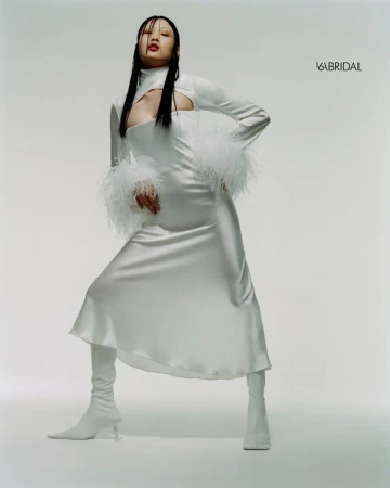 Asian woman poses in a white high-neck dress with feathered sleeves and matching boots against a pale background in a fashion shot.