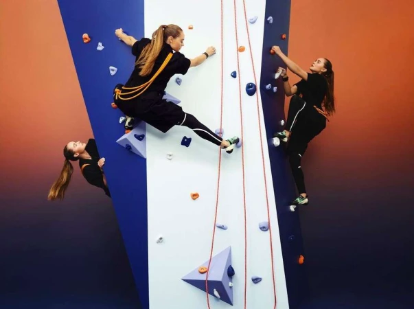 Three athletic women wearing sports attire are climbing an indoor rock climbing wall with blue and white sections, ropes, and colorful holds.