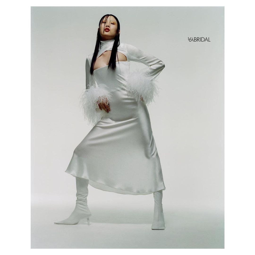 Model wearing a white cut-out dress with feathered cuffs and white boots against a neutral studio backdrop.