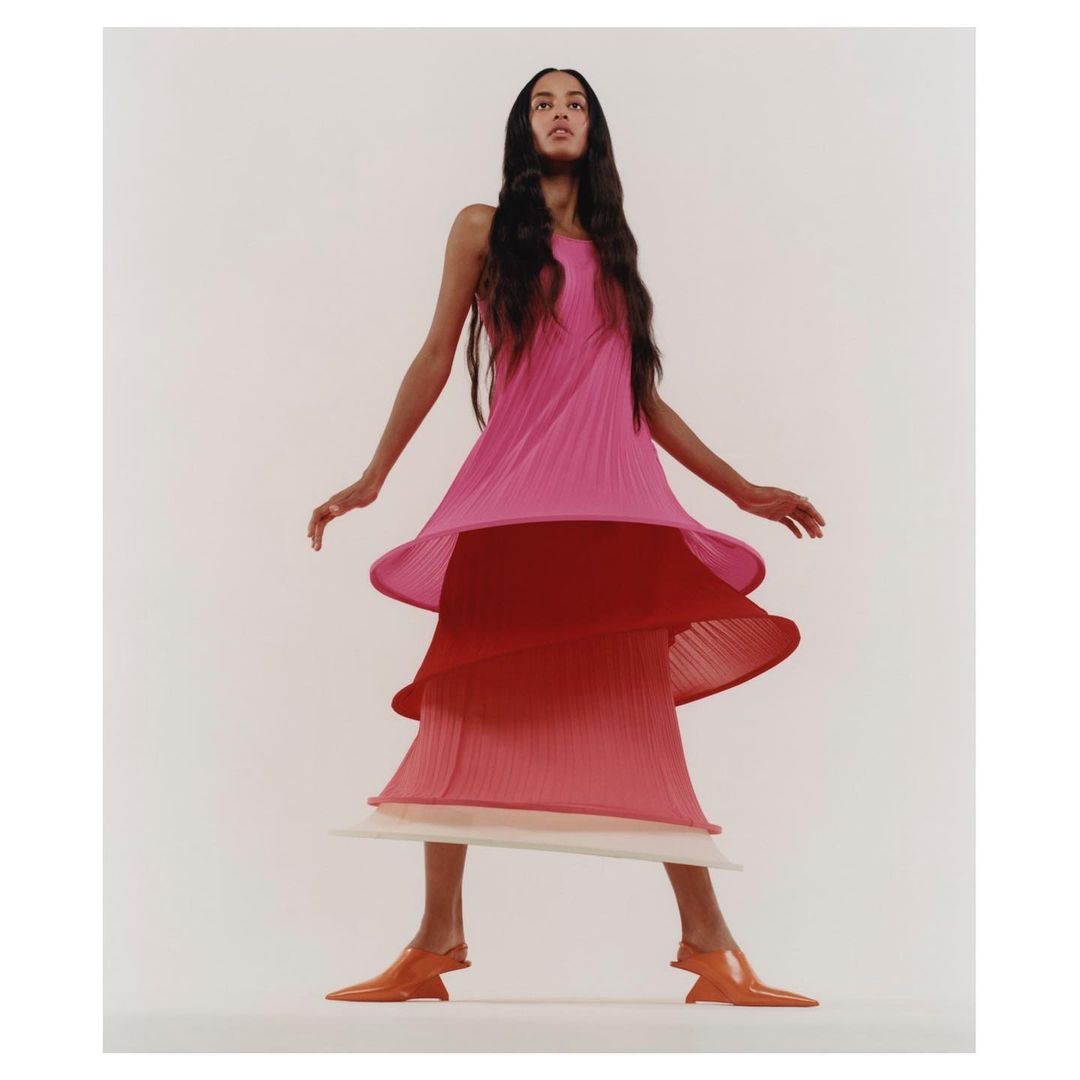 Fashion shot of a woman with long dark hair wearing a pink, red, and white tiered dress and orange shoes, posing against a white backdrop.