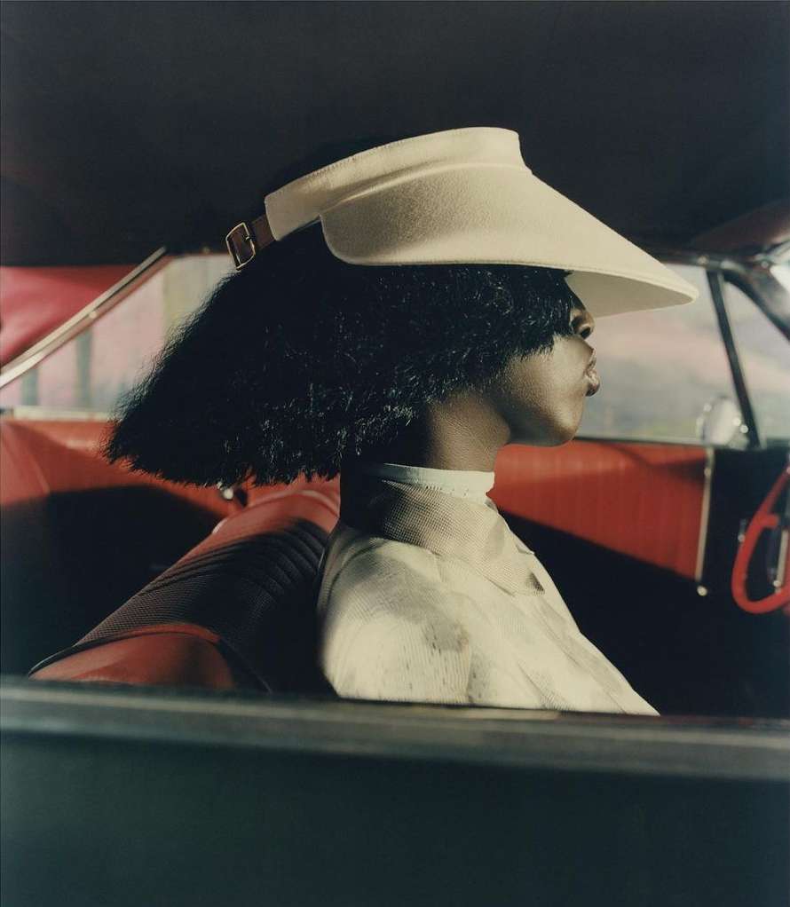 Elegant studio shot of a woman with dark hair wearing a visor in a vintage car with red interior and dark neutral background.