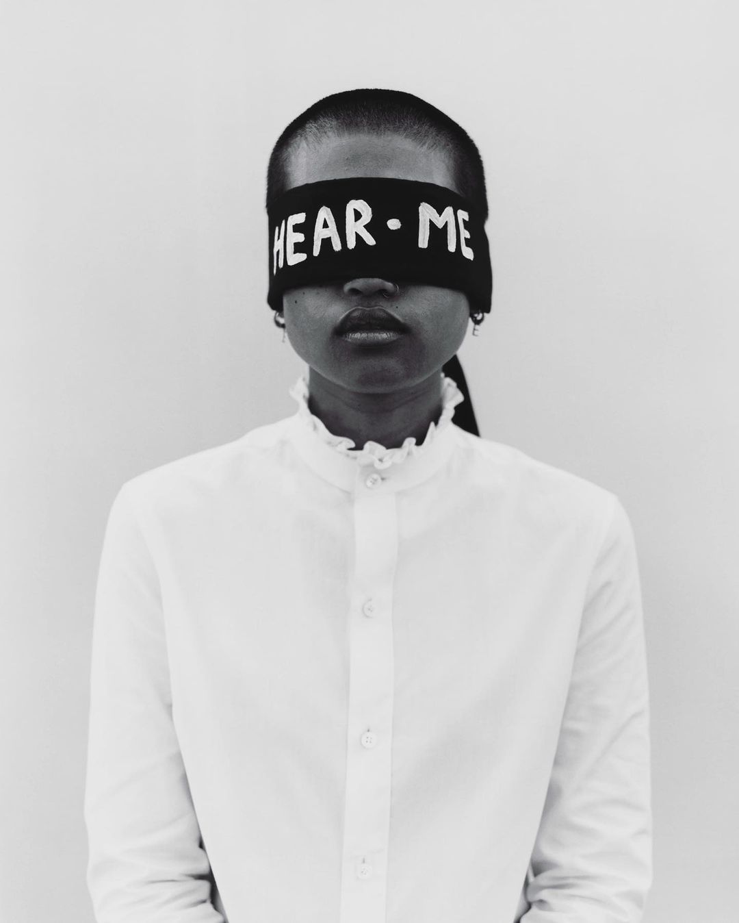 Close-up portrait of an adult woman with dark skin, wearing a white ruffled-collar blouse and a black headband with the text 'HEAR·ME'.