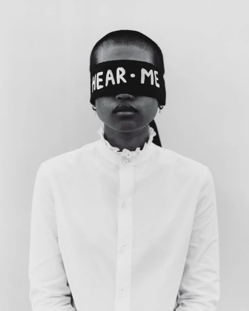 Close-up portrait of an adult woman with dark skin, wearing a white ruffled-collar blouse and a black headband with the text 'HEAR·ME'.