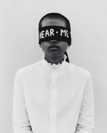 Close-up portrait of an adult woman with dark skin, wearing a white ruffled-collar blouse and a black headband with the text 'HEAR·ME'.