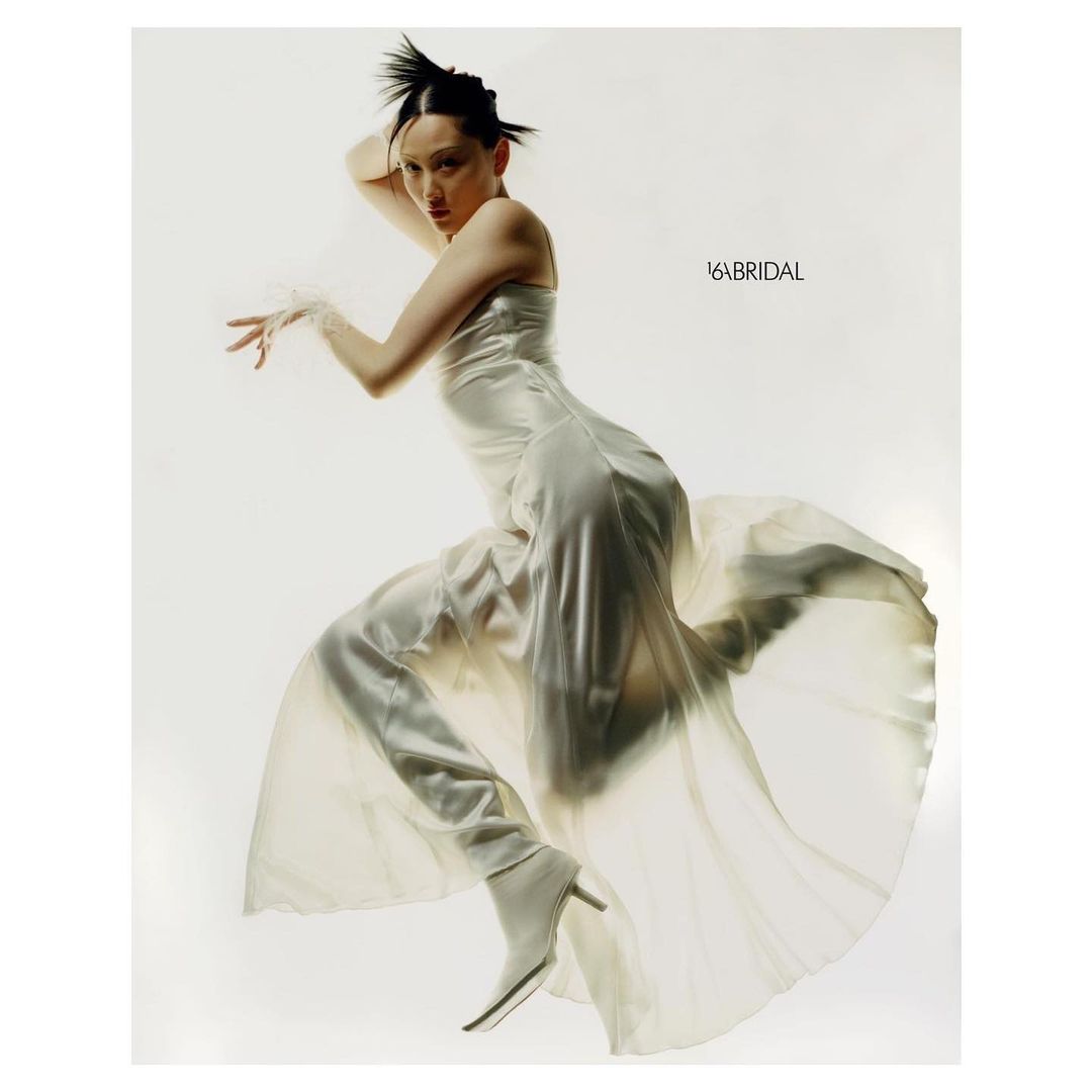 A woman in an off-white dress poses dynamically against a white backdrop, showcasing a modern bridal style with unique hair.
