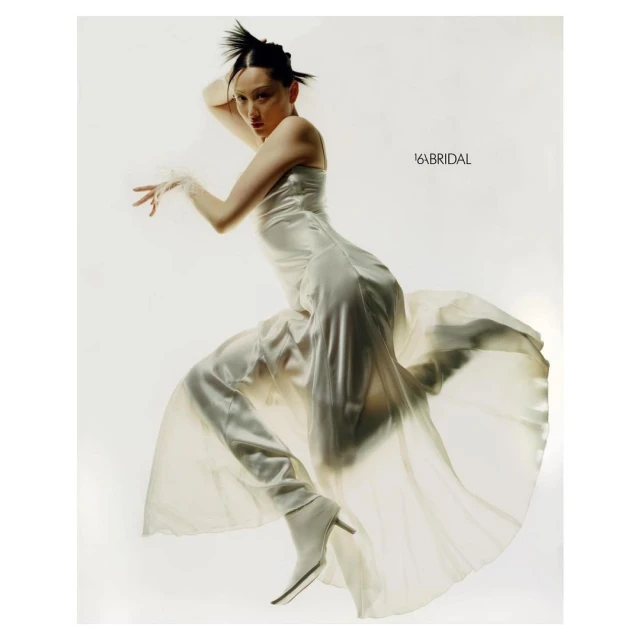 A woman in an off-white dress poses dynamically against a white backdrop, showcasing a modern bridal style with unique hair.