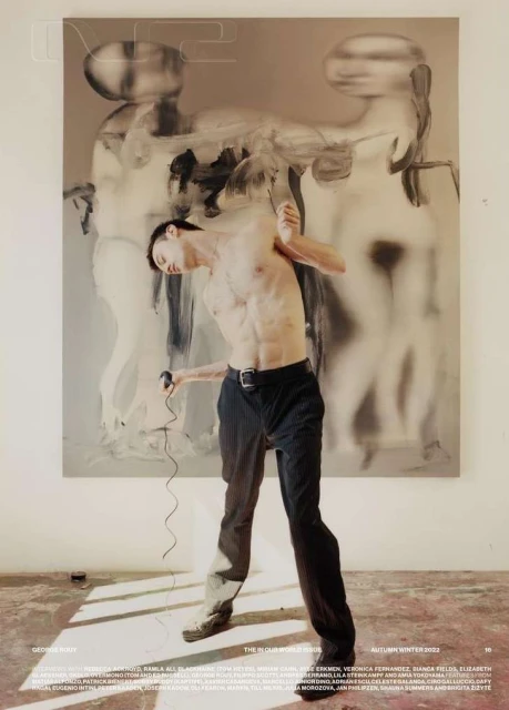 Fashion shot of a shirtless male model in black pants, standing in front of a large painting in a minimalist indoor setting.