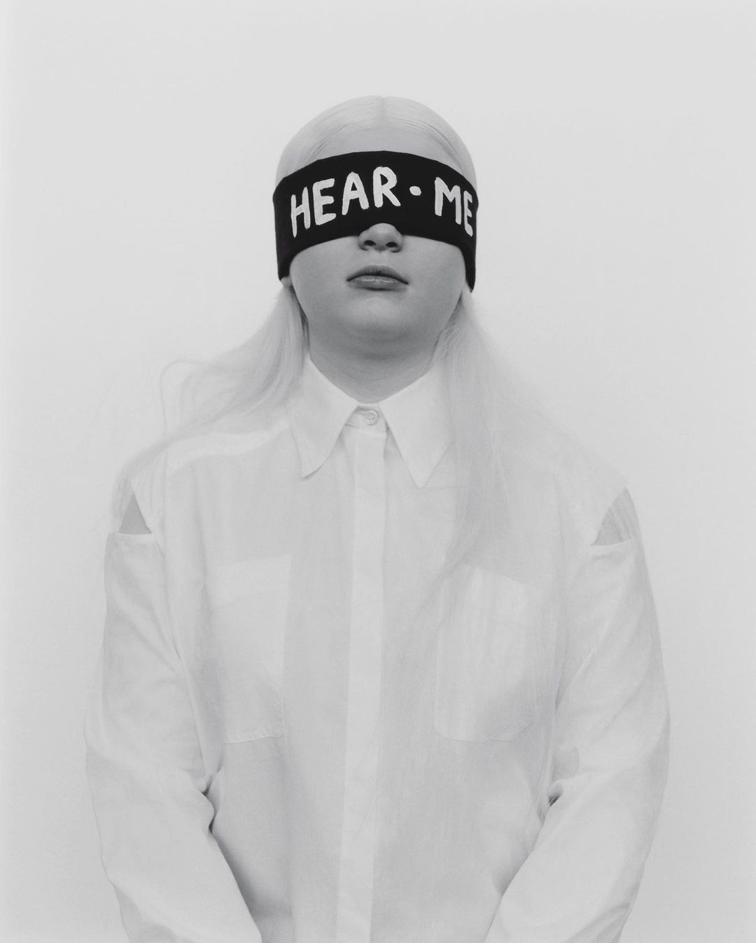 Portrait of a young adult with platinum blonde hair wearing a white button-down shirt and black blindfold with 'HEAR ME' in white letters.