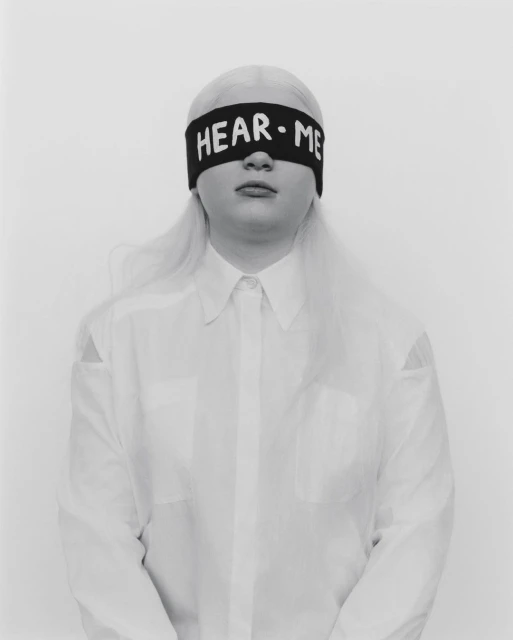 Portrait of a young adult with platinum blonde hair wearing a white button-down shirt and black blindfold with 'HEAR ME' in white letters.