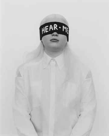 Portrait of a young adult with platinum blonde hair wearing a white button-down shirt and black blindfold with 'HEAR ME' in white letters.