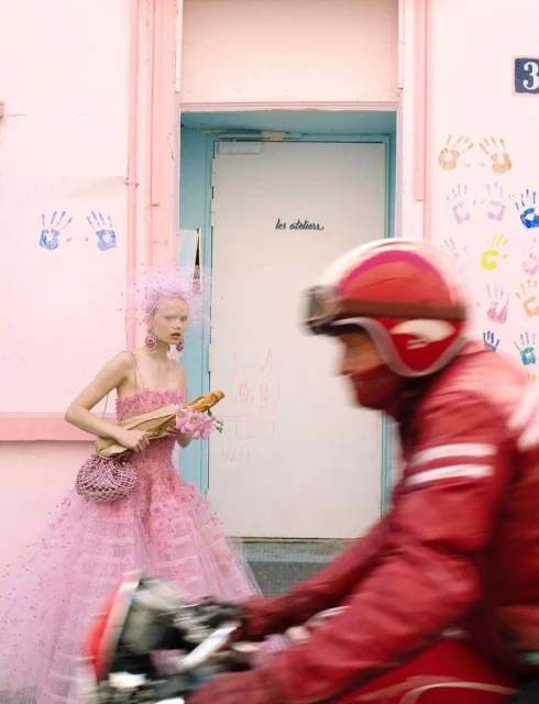 Fashion shot of a model in a pink dress holding flowers and bread while a person rides past on a red motorcycle. 