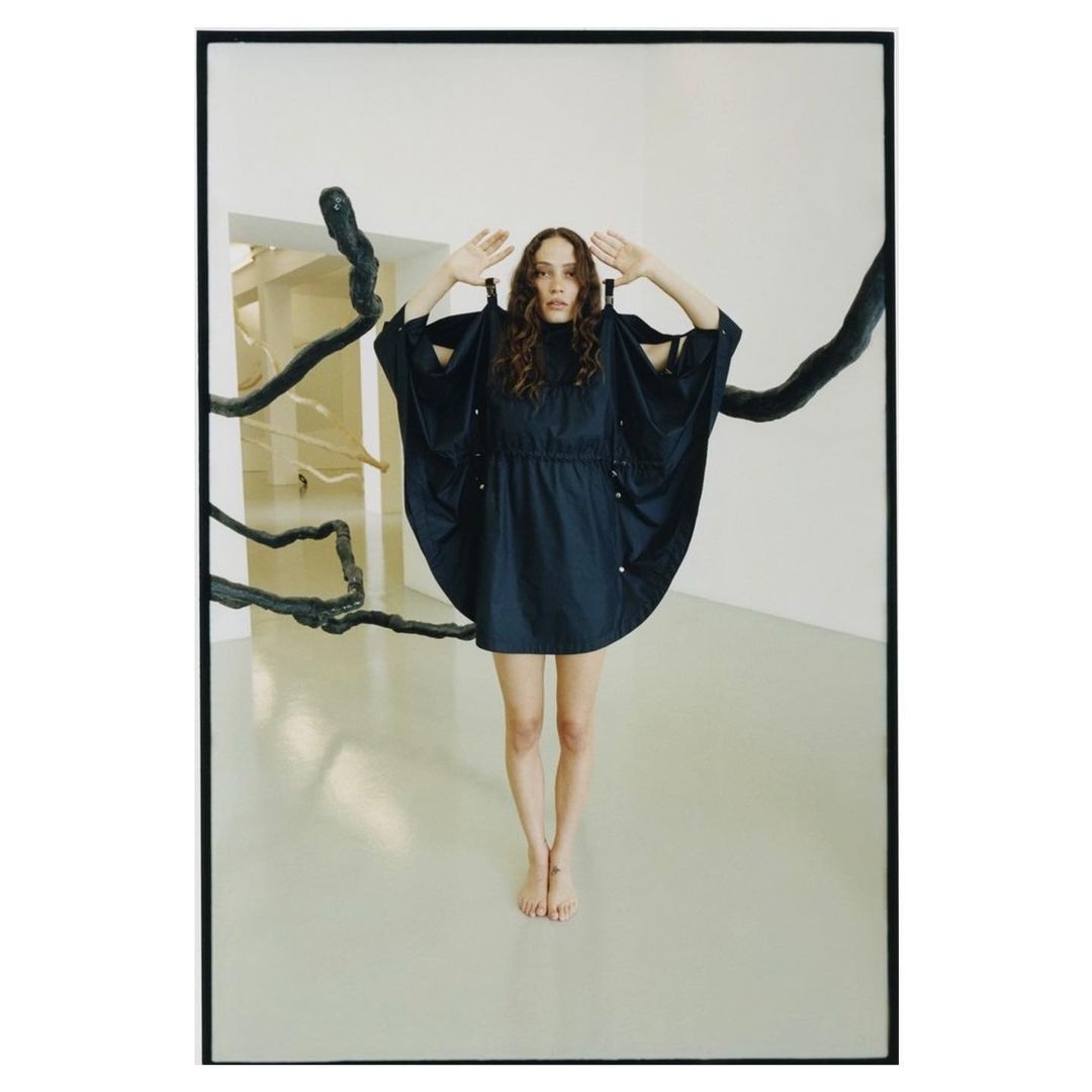 A woman in a short dark navy dress poses barefoot in a minimalist gallery with dark sculptural branches.