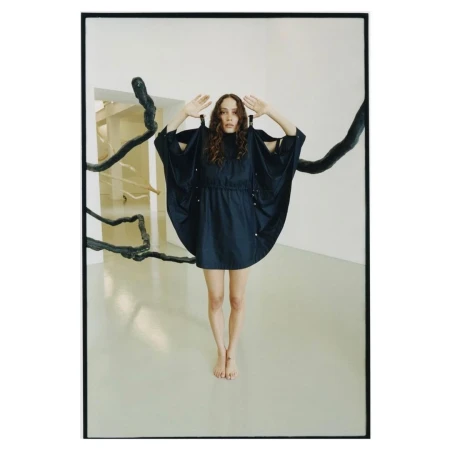 A woman in a short dark navy dress poses barefoot in a minimalist gallery with dark sculptural branches.