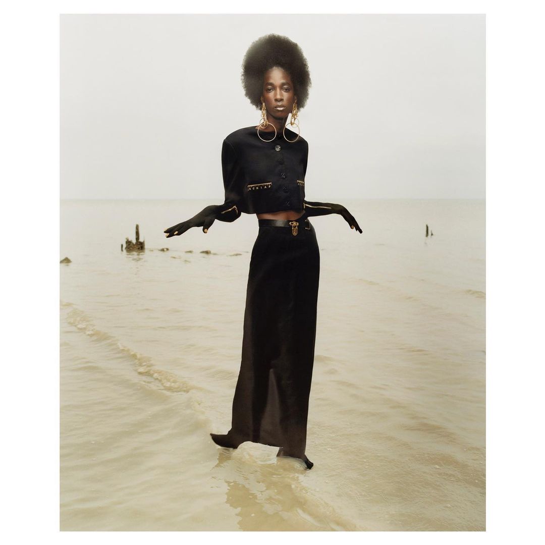 A lone Black woman in a black cropped jacket and a long skirt stands at the ocean's edge, exuding high fashion and poise against a muted sky.