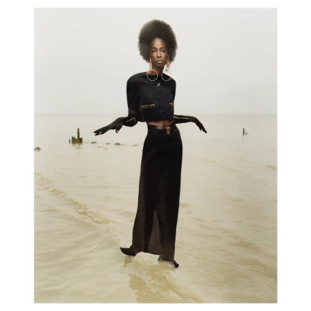 Model with large afro hairstyle wearing a black cropped jacket and long skirt standing in shallow water