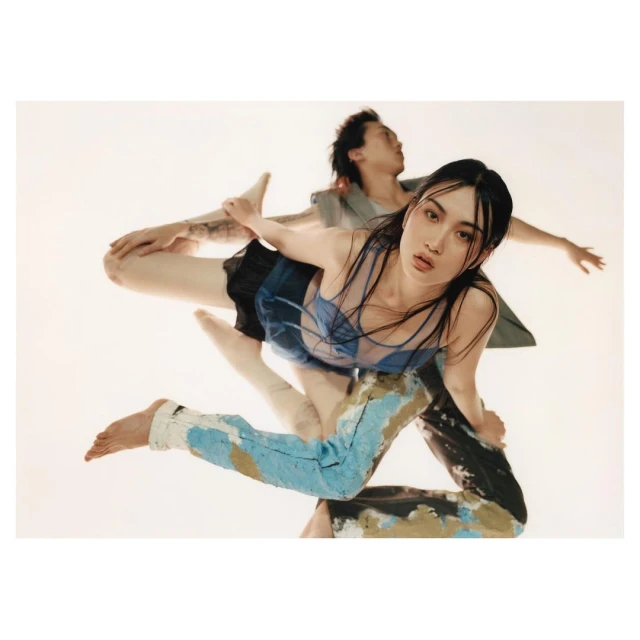 A woman in a sheer blue top and patterned jeans is supported by a man in a grey vest against a white backdrop in a studio shot.