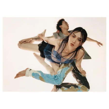A woman in a sheer blue top and patterned jeans is supported by a man in a grey vest against a white backdrop in a studio shot.