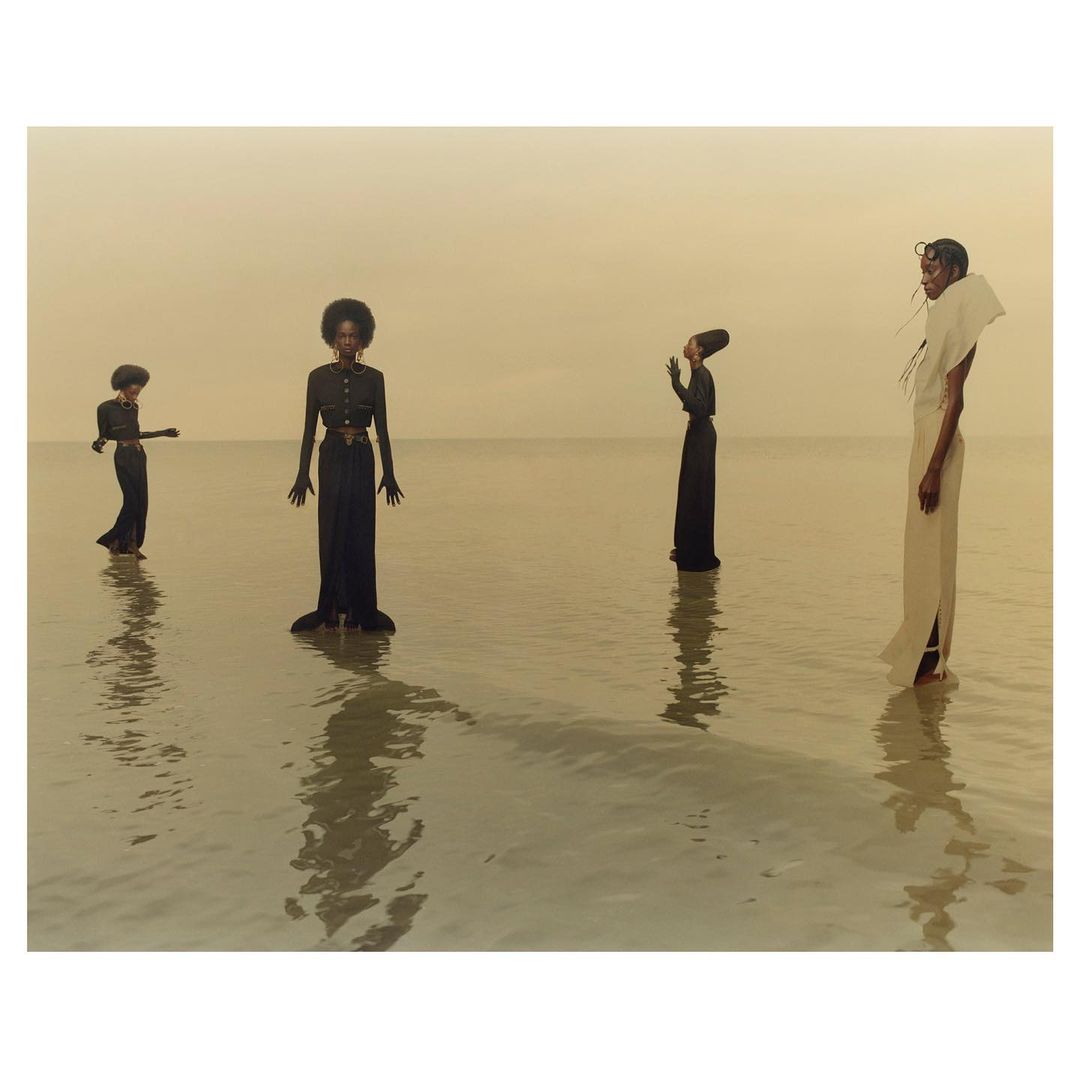 Four women stand in shallow water, wearing long gowns. The background features a calm, pale horizon with muted, earthy tones and subtle reflections.