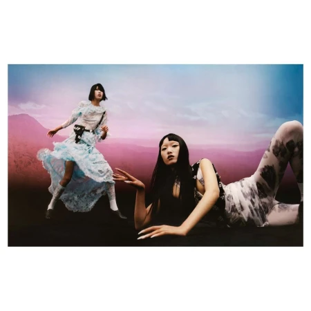 Two models with dark hair posing in a landscape setting with pink and blue gradient sky, one reclining and one standing