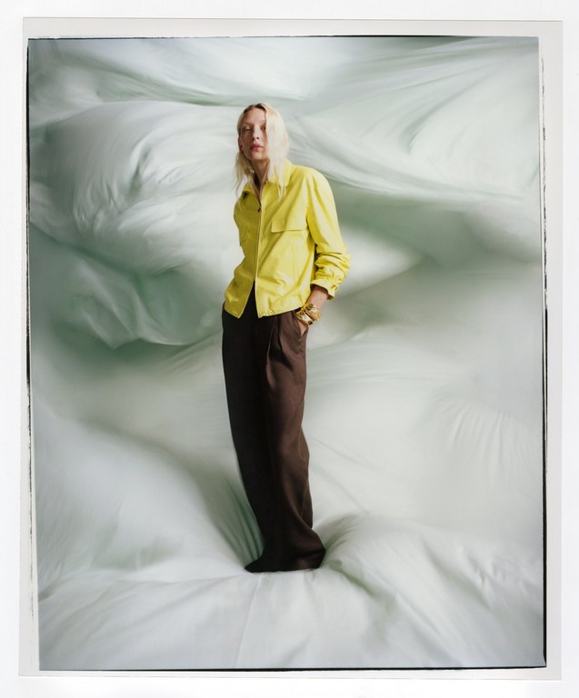 A stylish adult female poses in a yellow shirt and brown pants against a light-green background, full-length in a fashion-style photograph.