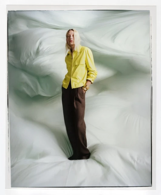 A stylish adult female poses in a yellow shirt and brown pants against a light-green background, full-length in a fashion-style photograph.